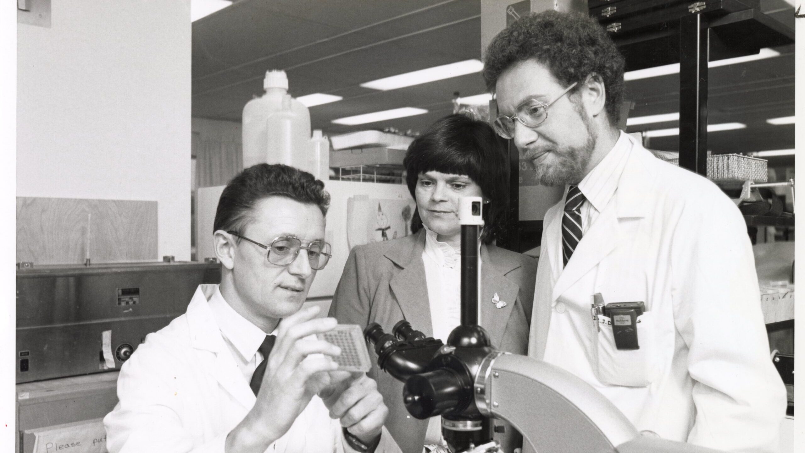 3 people in a lab, gathered around a microscope, in black and white.